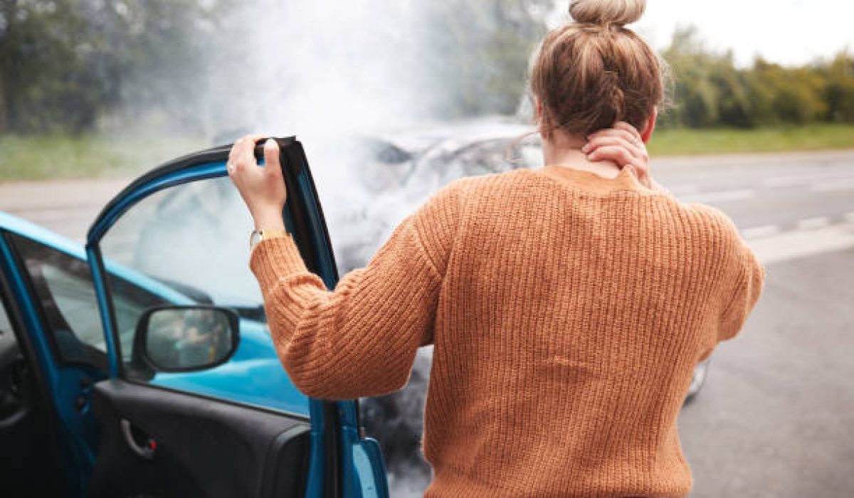 Rear View Of Female Motorist With Head Injury Getting Out Of Car After Crash